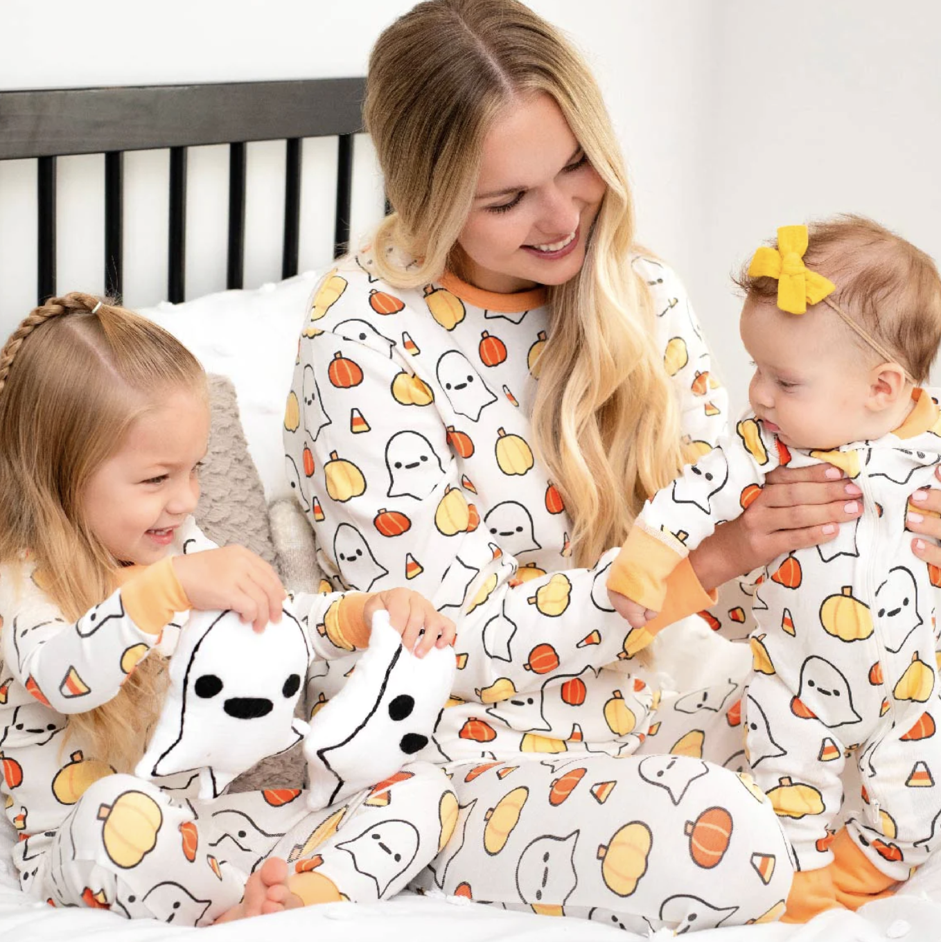 mom and toddler girl and baby girl in matching halloween pajamas. The print is white with ghosts, yellow and orange pumpkins, and candy corn with orange cuffs. The baby is wearing a yellow bow.  